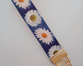 Könnte beinhalten: Blaues Schlüsselband mit Gänseblümchenmuster. Das Band zeigt weiße Blüten mit gelben Zentren und einen goldfarbenen Metallverschluss mit Schlüsselring. Der Hintergrund ist dunkelblau.
