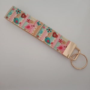 May include: A fabric wristlet key fob with a colorful patchwork design. The pattern includes pink, blue, and brown sections with bird, heart, and flower motifs. It has a gold-colored metal clasp and key ring.