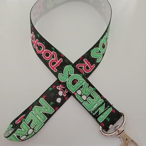 Può includere: Laccetto nero con la scritta "Nerds Rock" in verde e rosso. Il design include piccoli cuori e impronte di zampe. Un fermaglio in metallo color argento è attaccato all'estremità.
