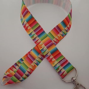 Puede incluir: Un cordón colorido con un patrón repetido de lápices de colores del arcoíris. El cordón tiene un cierre de metal plateado y está diseñado para usarse alrededor del cuello. Los lápices están dispuestos verticalmente, creando un diseño vibrante.