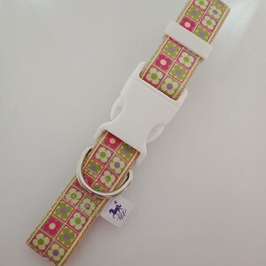 Puede incluir: Un collar para perros con hebilla de plástico blanco y anilla en D de metal plateado. El collar presenta un estampado floral con flores rosas, verdes y moradas sobre fondo beige. Lleva una pequeña etiqueta con un logotipo de caballo.