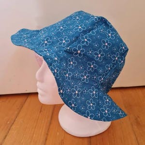 May include: A teal bucket hat featuring a repeating pattern of white-outlined flowers with small pink centres. The hat has a wide, downward-sloping brim and a rounded crown, displayed on a plain white mannequin head form.