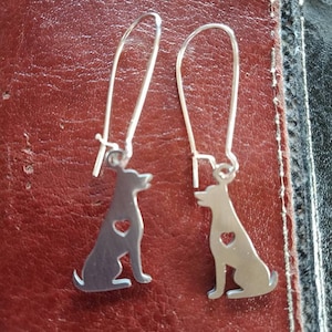 May include: A pair of silver dog-shaped earrings. Each earring has a sitting dog silhouette with a heart-shaped cutout. The earrings hang from long, curved ear wires. The earrings are displayed on a reddish-brown leather surface.