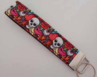 May include: A fabric wristlet key fob with a repeating pattern of skulls, hearts, roses, and eyeballs. The design features red, pink, white, and orange elements on a gray background. The key fob has a silver metal clasp and key ring.