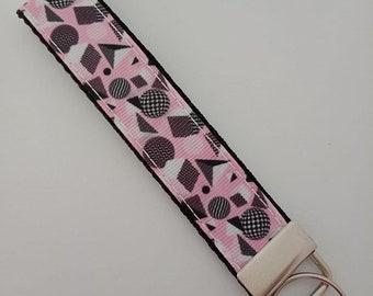 Könnte beinhalten: Ein Schlüsselanhänger mit rosa-weißem geometrischem Muster und schwarzem Rand. Das Muster umfasst Formen wie Kugeln, Dreiecke und Quadrate. Der Schlüsselanhänger hat einen silberfarbenen Metallverschluss und Schlüsselring.
