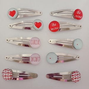 May include: A collection of silver metal hair clips with various designs. Some clips feature red hearts on a light blue background, while others have the words "Be mine," "XOXO," or "Love." The clips are likely for Valentine's Day or a similar occasion.