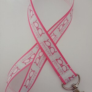 Puede incluir: Un cordón blanco con borde rosa y un patrón repetido de gafas y la palabra "Adorkable!". Tiene un cierre de metal plateado.