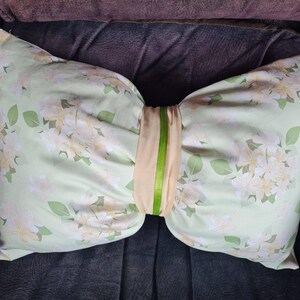 Peut inclure: Un oreiller décoratif en forme de nœud papillon. L'oreiller présente un motif floral vert clair avec des fleurs blanches et jaunes et des feuilles vertes. Une bande beige avec une rayure verte accentue le centre.