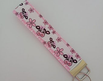 May include: A pink and white key fob with a cow and flower pattern. The key fob has a silver metal clasp and key ring. The cow design has a pink bow and the flowers are pink.
