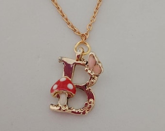 Könnte beinhalten: Goldfarbene Halskette mit einem Anhänger in Form des Buchstabens "B". Der Anhänger zeigt einen rot-weißen Pilz, einen rosa Schmetterling und dekorative Verzierungen. Die Kette ist ein filigranes Kettendesign.