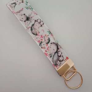 May include: A white wrist strap keyring featuring a repeating pattern of cartoon characters, flowers, and greenery. The keyring has a gold-coloured metal clasp and ring. The design features a mother and baby animal theme.