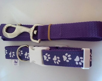 May include: A purple dog collar and leash set. The collar and leash are made of a woven material. The collar has a white plastic buckle and a pattern of white paw prints. The leash has a silver metal clasp.