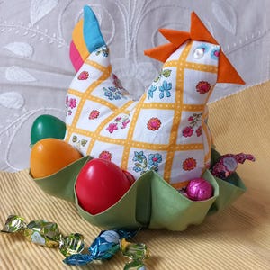 Può includere: Una gallina di stoffa gialla e bianca con una coda verde, arancione e blu. La gallina tiene un nido di stoffa verde con quattro uova colorate al suo interno. Ci sono anche diversi dolci avvolti sul tavolo.