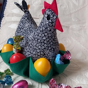 Può includere: Una gallina di stoffa con motivo floreale bianco e nero, con cresta e becco rossi. La gallina tiene un nido di stoffa verde con uova di Pasqua colorate e caramelle avvolte.