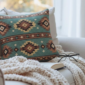 May include: A decorative throw pillow with a teal background and a geometric pattern in brown, tan, and black. The pillow is on a cream-colored sofa with a matching blanket, a pair of glasses, and an open book.
