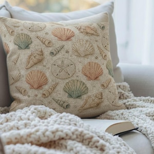 Puede incluir: Un cojín cuadrado beige con un patrón de conchas marinas en tonos melocotón, verde y beige. El cojín está sobre un sofá blanco con una manta de punto crema y un libro abierto. El diseño incluye varias formas de conchas marinas.