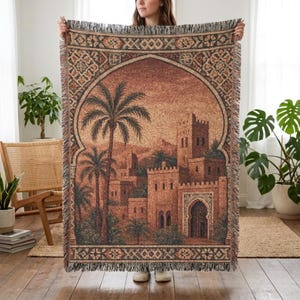 Könnte beinhalten: Gewebte Tapeten-Wurfdecke mit einer detaillierten Illustration einer nahöstlichen Stadt mit Palmen und einer Festung. Die Decke hat eine braune und beige Farbpalette mit dekorativen Fransenrändern. Das Design wird von einem Torbogen umrahmt.
