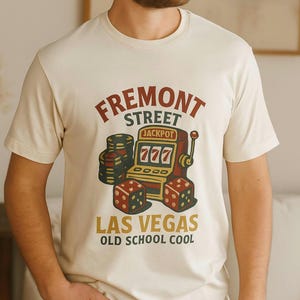 May include: Cream-colored t-shirt featuring a vintage-style graphic. The design includes the words "FREMONT STREET LAS VEGAS" with a jackpot-themed slot machine, dice, and the phrase "OLD SCHOOL COOL".