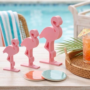 May include: Three pink flamingo figurines of varying heights stand on rectangular bases. Beside them, a round woven rattan tray holds a glass of orange liquid with a lemon slice and cocktail umbrella, a stack of napkins, and a gold spoon.