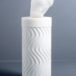 May include: A white tissue box with a cylindrical shape and a wavy, textured pattern. The top has a textured band and a tissue is pulled out. The tissue box is on a reflective surface.