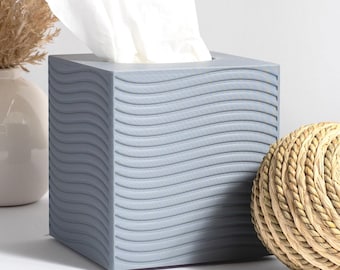 Könnte beinhalten: Grauer Tissuebox-Bezug mit wellenförmigem Design. Der Bezug ist aus strukturiertem Material und hat eine quadratische Form. Die Tissuebox steht auf einer weißen Oberfläche neben einer Vase mit Trockenblumen und einem geflochtenen Ball.