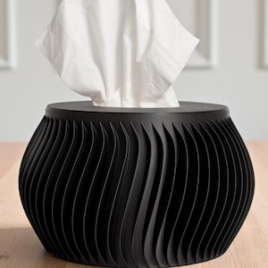 May include: A black tissue box with a unique, wave-like design. White tissues are visible, emerging from the top opening. The box sits on a light-colored wooden surface, with a white wall in the background.