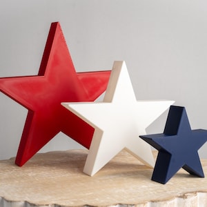 May include: Three wooden stars in red, white, and blue, arranged in a decreasing size order on a light brown wooden surface.