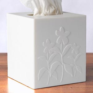 May include: White ceramic tissue box cover with an embossed floral design. The cover is square and has a slot for tissues to be pulled out.