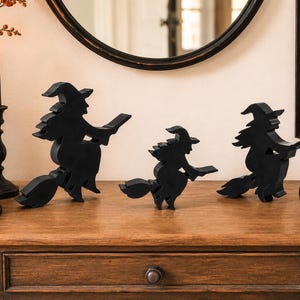 May include: Three black wooden witch silhouettes in varying sizes, each depicted riding a broomstick. The figures are solid black, featuring pointed hats and flowing robes, designed as freestanding decorative items for a flat surface display.