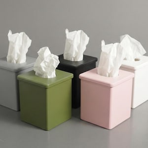 May include: A collection of six square tissue box covers in various colors: gray, black, green, pink, and white. Each box has a square opening at the top with white tissues protruding.