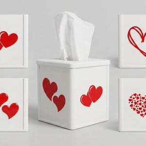 May include: A white tissue box with red heart designs. The box has a square shape with a removable top. Several small white boxes with red heart designs surround the tissue box.