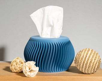 Könnte beinhalten: Eine blaue Taschentuchbox mit wellenförmigem Design, die weiße Taschentücher enthält. Dekorative Kugeln aus natürlichen Materialien liegen auf einer Holzoberfläche. Ein modernes Wohnaccessoire.