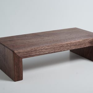 May include: A rectangular wooden riser with a dark brown finish and visible wood grain. The stand features a flat top surface supported by two vertical side panels, creating a bridge-like structure with a clean, simple, and functional design.