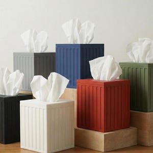 Puede incluir: Una colección de coloridas cajas de pañuelos en negro, blanco, azul, rojo, gris y verde. Cada caja es cuadrada con detalles de costillas verticales y contiene pañuelos blancos.