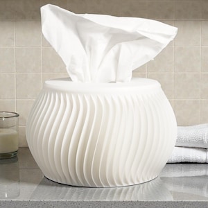 May include: A white tissue box with a swirling, textured design. The tissue box is round and sits on a countertop next to a glass, a soap dispenser, and folded white towels. White tissues are pulled from the top.