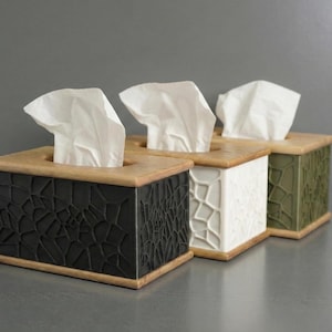 Peut inclure: Trois boîtes à mouchoirs décoratives avec des dessus en bois et des côtés à motifs. Les boîtes sont noires, blanches et vert olive. Chaque boîte contient des mouchoirs blancs sortant du haut. Les boîtes sont alignées.