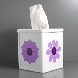 May include: White tissue box with two purple daisy flower designs. The tissue box is square and has a lid. White tissues are pulled from the top of the box. The background is a solid gray color.