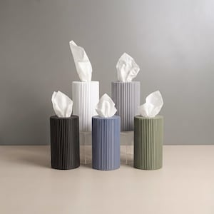 May include: A collection of five cylindrical tissue holders featuring a vertical ribbed texture. The holders are available in black, white, gray, blue, and olive green, each containing a white tissue extending from the top opening.