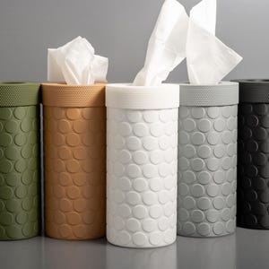 May include: Five cylindrical tissue box holders in olive green, tan, white, grey, and black. Each has a textured, dotted pattern and a matching lid. White tissues are visible in some.