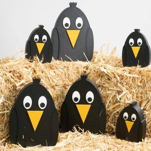 May include: A collection of six black wooden bird figurines with white eyes and yellow beaks, arranged on a bed of hay. The birds vary in size, creating a whimsical display. The background is a plain white.