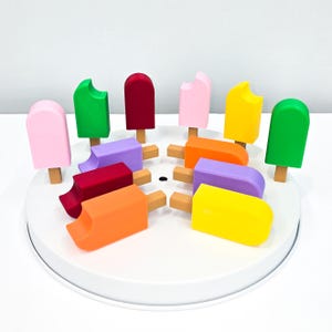 May include: A collection of colorful wooden popsicle toys arranged on a white circular base. The popsicles are in various colors, including pink, green, red, purple, orange, and yellow, with some showing bite marks. Each popsicle has a wooden stick.