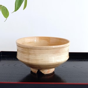 May include: A light brown ceramic tea bowl with a rounded shape and three small feet. The bowl is sitting on a black tray with a red border.