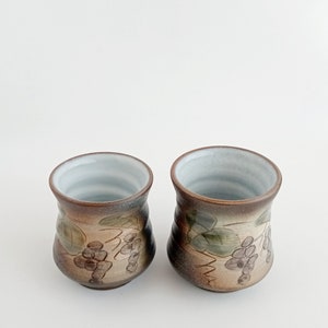 Puede incluir: Dos tazas de cerámica marrón con un esmalte blanco y un diseño de vid. Las tazas tienen un fondo redondeado y una parte superior ligeramente acampanada.