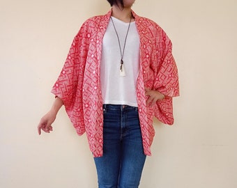 Puede incluir: Una chaqueta estilo kimono con estampado rojo y blanco con mangas anchas. La chaqueta se lleva sobre una camiseta blanca y vaqueros azules. El estampado es un diseño repetitivo de pequeñas formas blancas sobre fondo rojo.