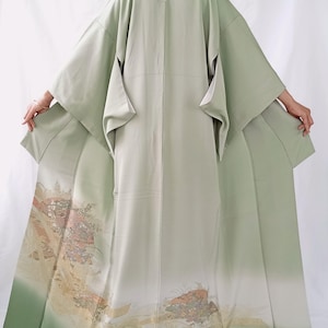 May include: A sage green kimono featuring wide sleeves and a floor-length hem. The lower portion of the garment displays a detailed landscape illustration in muted tones of gold, brown, and blue, depicting stylised foliage and architectural elements.