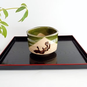 May include: A green and brown ceramic teacup with a stylised fox design. The cup is sitting on a black tray with a red border.
