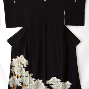 May include: A black kimono featuring wide sleeves and three small circular crests along the upper back. The lower portion of the garment is decorated with an embroidered pattern of light blue and white pine trees with tan trunks.