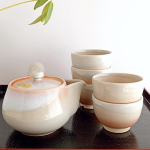 Op de afbeelding: Een crèmekleurige keramische theeservies, bestaande uit een theepot en vier bijpassende kopjes. De theepot heeft een afgeronde vorm, een deksel en een tuit. De kopjes en de theepot hebben een perzikverloop aan de basis. De set staat op een zwarte dienblad.