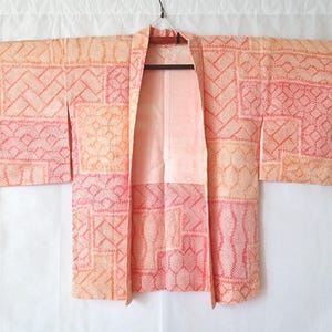 May include: A peach and coral colored kimono jacket featuring a patchwork design of various geometric patterns including chevrons, diamonds, and grid lines. The garment has wide sleeves, an open front, and a light pink interior lining.