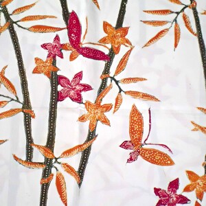 May include: White fabric with a repeating pattern of orange and brown leaves, pink and red flowers, and orange and red butterflies. The pattern is reminiscent of a traditional Indonesian batik design.